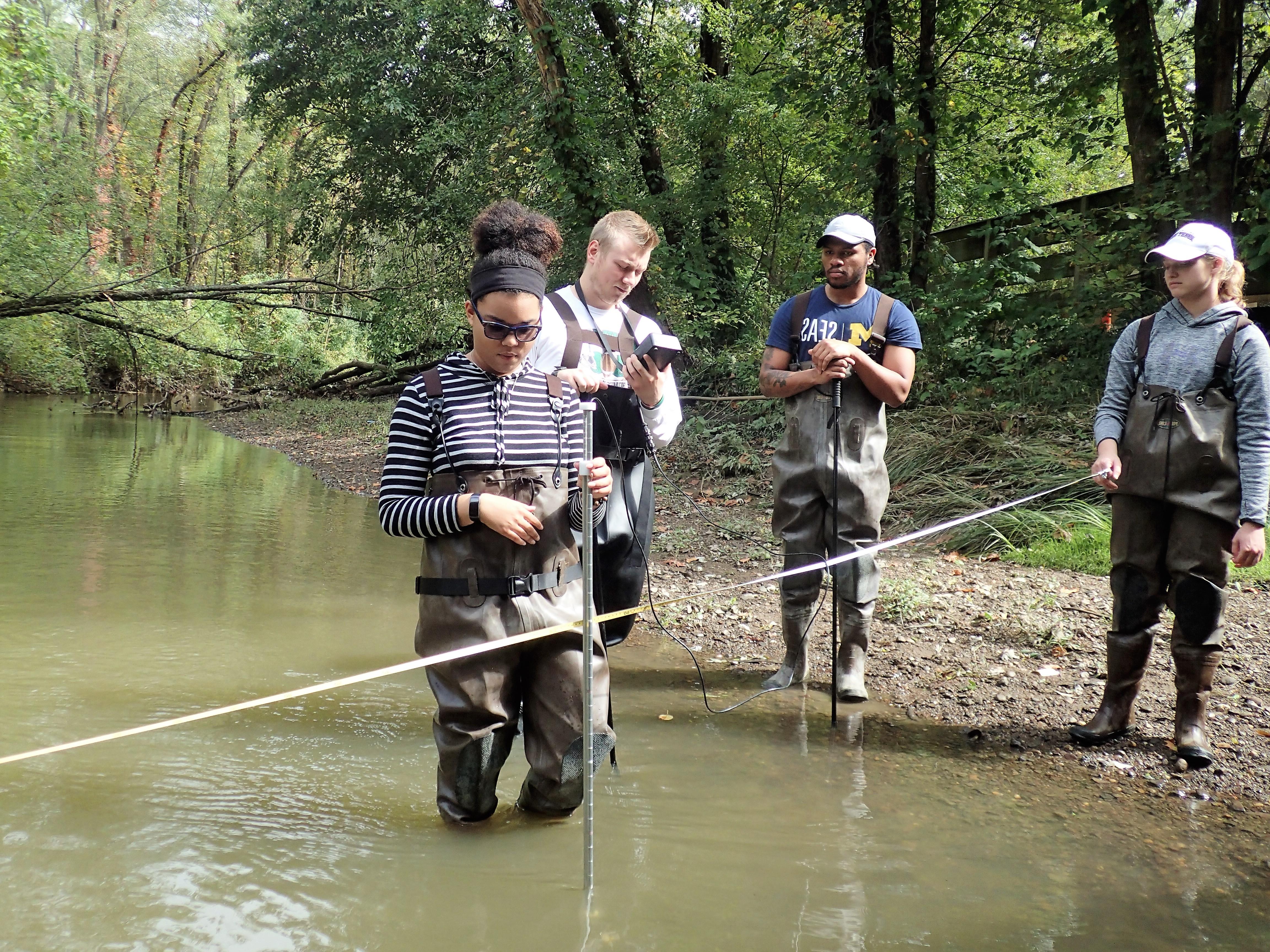 Four environmental science students wade in the water with a rope and measurement tools to evaluate river flow